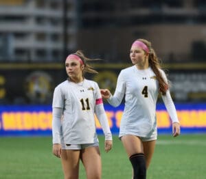 Two female soccer players in white jerseys and pink headbands on the field, numbers 11 and 4 visible.
