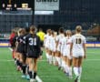Two soccer teams walk onto the field before a match, one in black and the other in white.