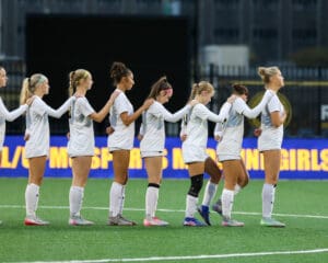 Women's soccer team lined up in unity on field, holding each other's shoulders before a match.