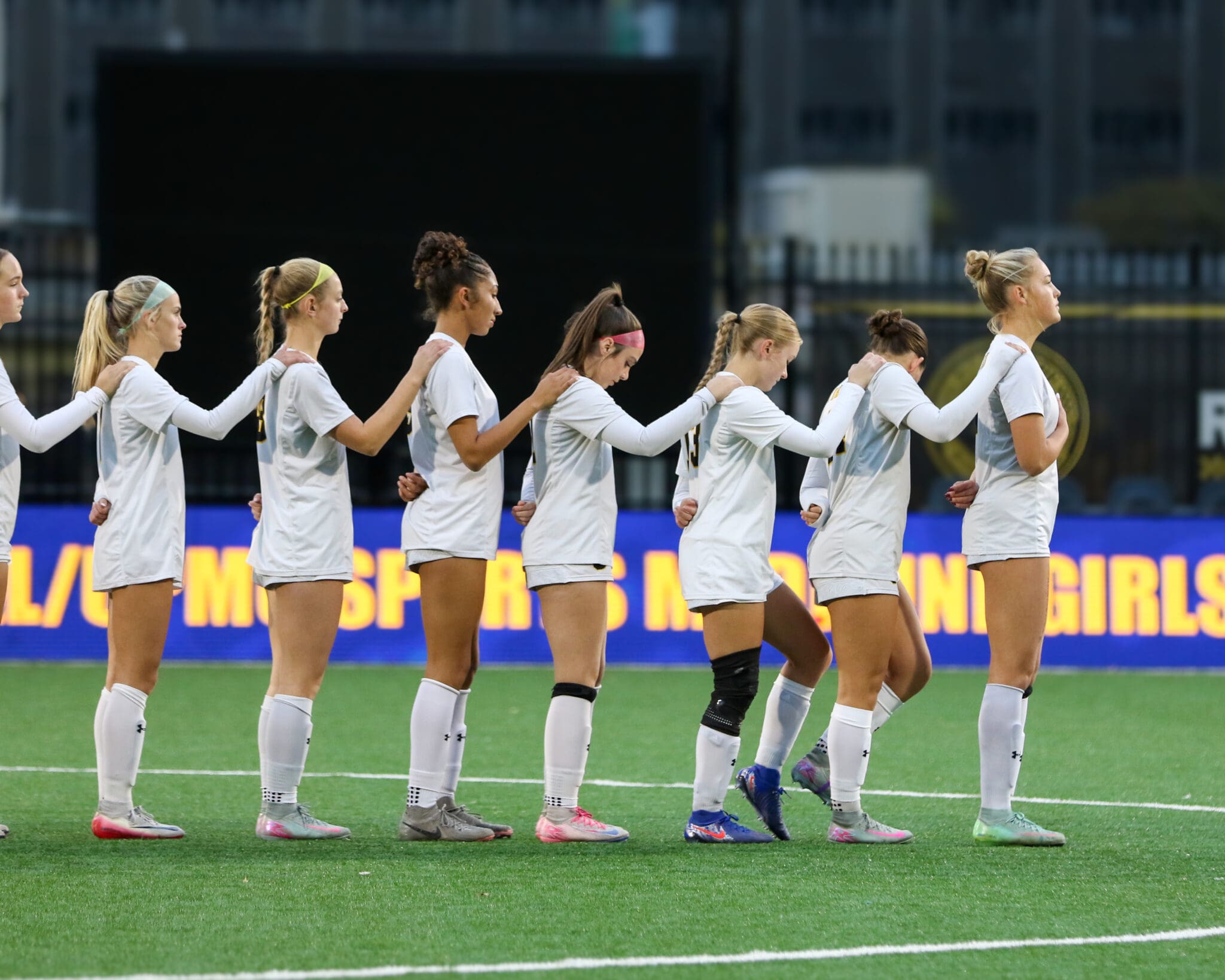 Women's soccer team lined up in unity on field, holding each other's shoulders before a match.