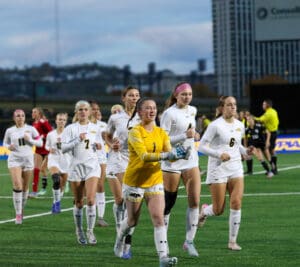 Women's soccer team in white uniforms warming up on the field before a match, with a goalie in yellow leading.