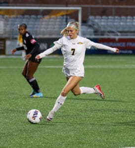 Soccer player in white uniform kicking ball during a match on a green field, with focused action and determination.