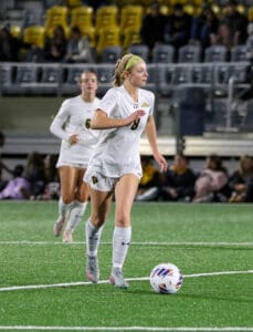 Soccer player in white uniform dribbling ball during a night match on a green field.
