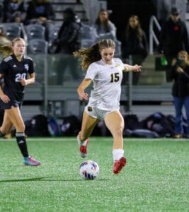 Soccer player in white jersey dribbles ball on field during night match, with spectators in the background.