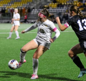 Soccer player in white uniform dribbles ball against opponent in black jersey on a green field at night.