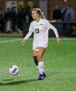 Soccer player in white uniform dribbling a ball on a green field during a match at night.