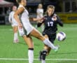 Two female soccer players vie for the ball on a brightly lit field during a competitive match.