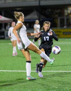 Two female soccer players vie for the ball on a brightly lit field during a competitive match.