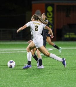 Soccer player in white jersey number 2 dribbles past opponents during nighttime match on green field.