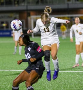 Soccer players compete for a header during a night match on a vibrant green field, intense action captured mid-air.