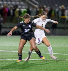 Two female soccer players competing intensely for the ball on a field during a match.
