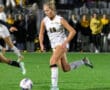 Female soccer player in white uniform dribbles ball during night match on grass field.