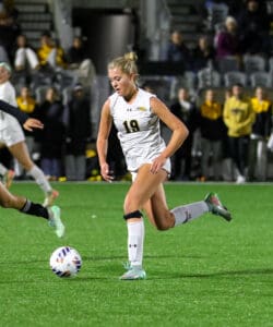 Female soccer player in white uniform dribbles ball during night match on grass field.
