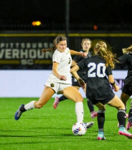Women's soccer match at night, player in white dribbling ball against opponent in black, on green field.