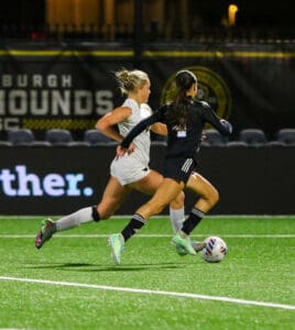 Two female soccer players competing for the ball on a well-lit stadium field at night.