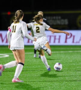 Two female soccer players in white uniforms running and kicking a ball on a green field during a nighttime match.