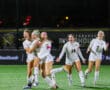 Soccer team celebrates a goal on the field at night, players in white jerseys, joyful expressions.