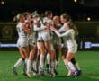 Women's soccer team celebrates a victory with a group hug on the field at night.