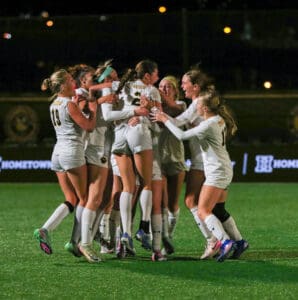 Women's soccer team celebrates a victory with a group hug on the field at night.