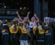 Girls' soccer team celebrating victory on field at night, wearing matching jackets, arms raised in joy.