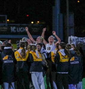 Girls' soccer team celebrating victory on field at night, wearing matching jackets, arms raised in joy.