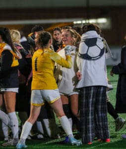 Soccer team celebrates victory on field, players in yellow and white uniforms joyfully embrace under night lights.