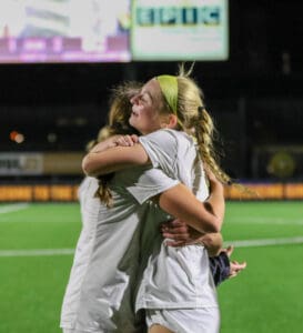 Two female soccer players joyfully embrace on the field, celebrating a victory under the stadium lights.