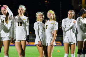 Female soccer team players standing in line on a nighttime field, wearing matching white uniforms.