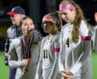 Three female soccer players in white jerseys, with medals, celebrating on the field at night.
