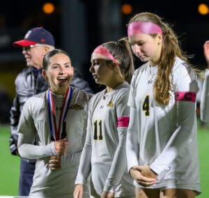 Three female soccer players in white jerseys, with medals, celebrating on the field at night.