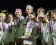 Victorious girls' soccer team celebrates with championship trophy and medals at night.