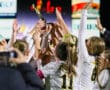 Girls soccer team celebrates with trophy under stadium lights. Teamwork and victory in sports event.