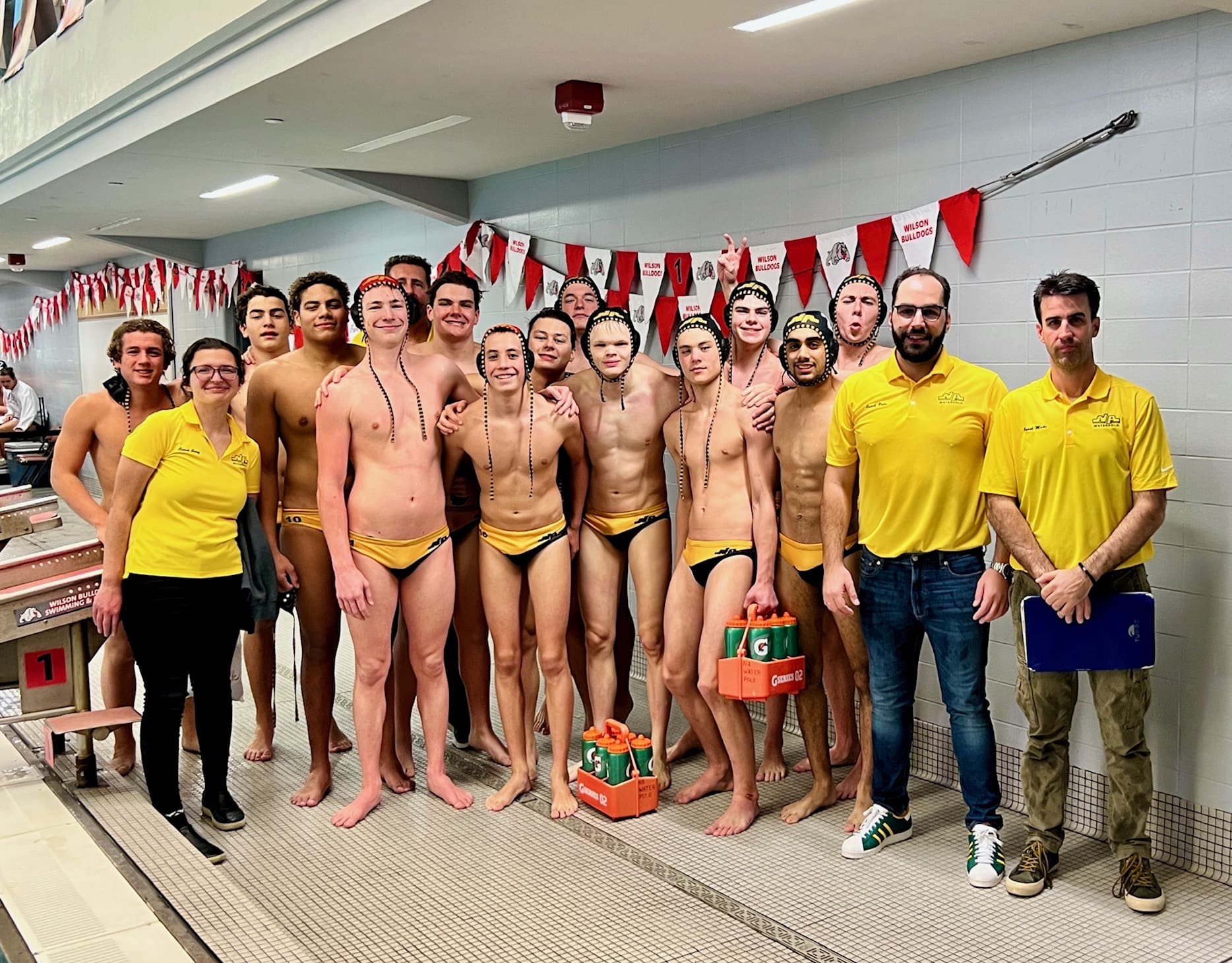 Water polo team posing in swimsuits and helmets by the poolside, with three coaches in yellow shirts.