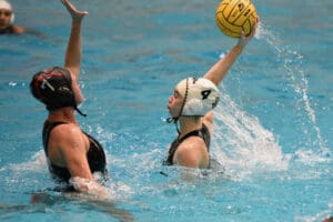 Two water polo players in action, one reaching high with the ball, intense gameplay in a pool setting.