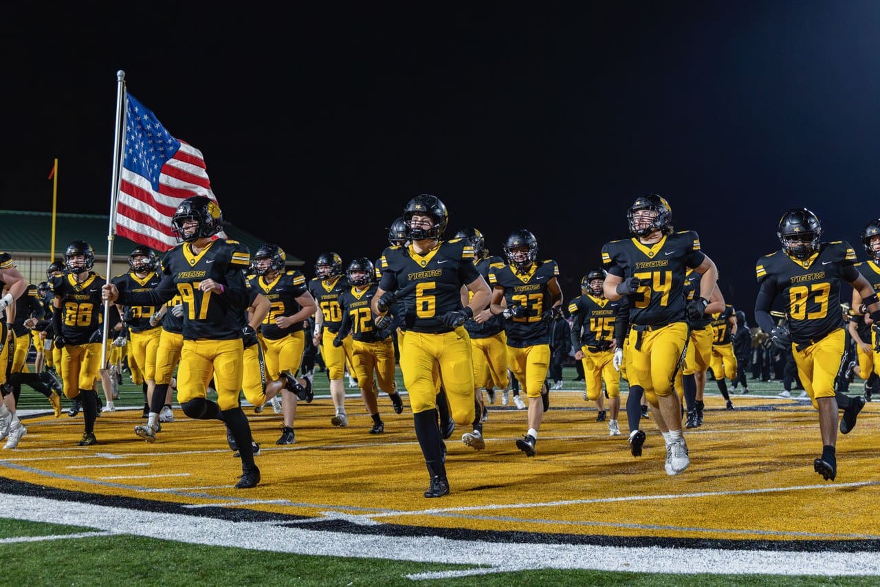High school football team in black and yellow uniforms running onto the field, carrying the American flag.