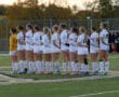 Girls' soccer team lined up on field, wearing white uniforms with numbers, ready for a match at sunrise.