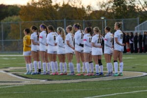 Girls' soccer team lined up on field, wearing white uniforms with numbers, ready for a match at sunrise.