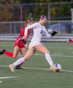 Soccer player in white uniform dribbling ball on field with opponent in red, action-packed sports scene.