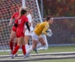 Goalkeeper in yellow jersey secures soccer ball during a match, surrounded by players in red and white uniforms.