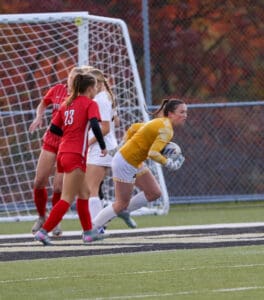 Goalkeeper in yellow jersey secures soccer ball during a match, surrounded by players in red and white uniforms.