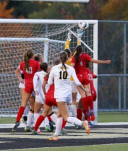 Soccer goalkeeper jumps to block a shot during a match, players in action near the goalpost.