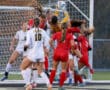 Soccer goalie catches ball during intense match near goal, surrounded by players in red and white uniforms.