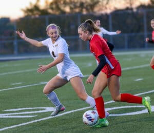 Two soccer players compete for the ball on a field during a match at sunset.