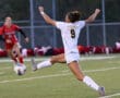 Soccer player in white uniform mid-kick on field, opponent approaching in red; intense match action.