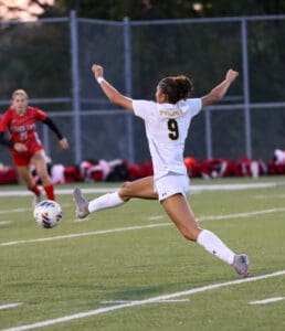 Soccer player in white uniform mid-kick on field, opponent approaching in red; intense match action.