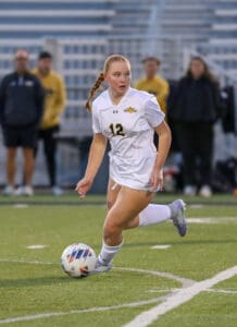 Soccer player in white jersey dribbling the ball on a green field during a match.