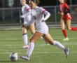 Soccer player in white uniform dribbles ball during a night match on a grass field with focused teammates nearby.