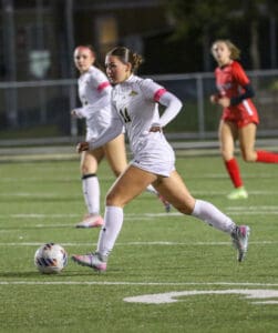 Soccer player in white uniform dribbles ball during a night match on a grass field with focused teammates nearby.