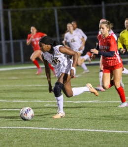 Soccer player dribbling the ball during a night game, with opponents in pursuit on the field.