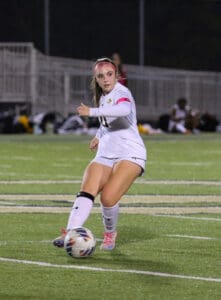Soccer player in action on the field, wearing a white uniform and pink headband, focused on the ball.